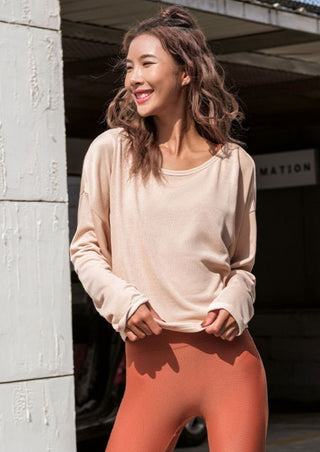 A woman with partially tied wavy hair smiles outdoors, wearing an apricot Take it All In Sweater and high-waisted orange leggings. She stands near a light wall with part of a building in the background.