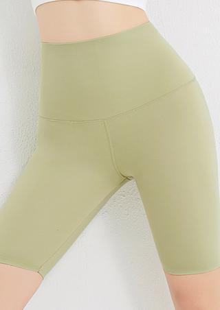 Someone wearing Voguish Compact Yoga Shorts in Grass Green and a white crop top stands against a textured white wall, with the image highlighting the shorts' fit and design on the lower torso and upper thighs.