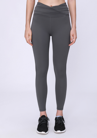 A person in PE High Rise Tight Pants, gray with a criss-cross waistband, stands against a plain background. Their hands rest at their sides, and they wear black athletic sneakers with white soles. The image centers on the lower body, excluding the face.
