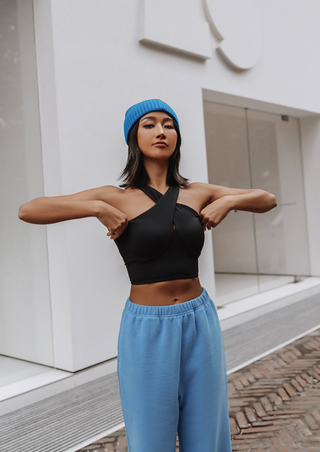 A woman stands confidently outside a modern building with glass doors, wearing a black Studio Lite Halter Tube, blue beanie, and blue sweatpants. Her straight hair flows loosely as she stands on the cobblestone in front of the minimalist white facade.