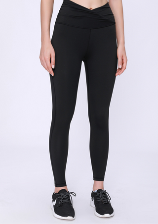 A person stands against a plain white background in PE High Rise Tight Pants, Classic Black, and black-white athletic shoes. The leggings have a wrap-style waistband. Only the lower torso and legs are visible, with the right hand resting by their side.