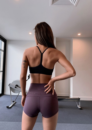 A woman with long hair and a palm tree tattoo on her left arm is in a workout room, wearing a Unity Racerback Sports Bra and purple shorts. Exercise equipment like a stationary rower is visible, along with large windows and ceiling lights.