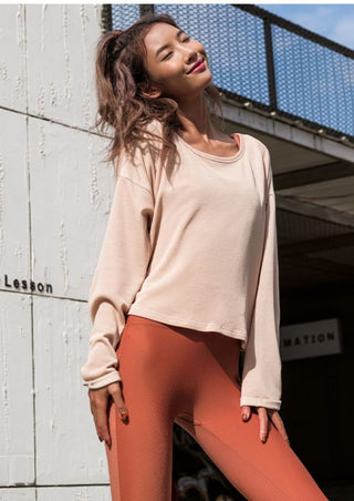 A woman wearing the Take it All In Sweater and orange leggings smiling with closed eyes stands in front of a concrete wall and building with a metal railing. Her hair is tied up, and the clear sky suggests it's a sunny day.