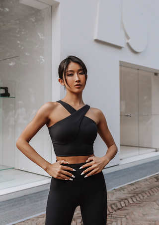 A woman confidently stands before a modern building, wearing a Studio Lite Halter Tube and leggings. Her hair is tied back, and her hands rest on her hips as glass and white walls provide a minimalist backdrop.