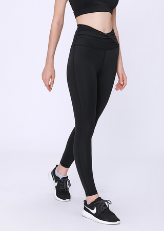 A person stands against a plain backdrop wearing a black sports outfit. They're in a black sports bra paired with PE High Rise Tight Pants and black sneakers featuring white soles. Their stance is casual and relaxed, one foot slightly ahead of the other.