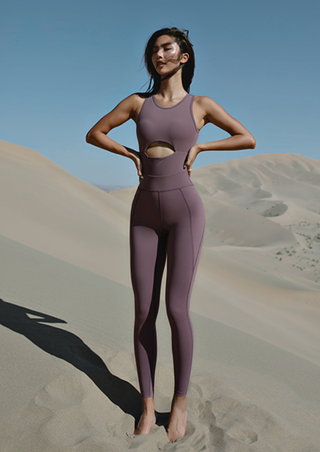 A person stands barefoot on sand dunes, wearing the Cutout Freeform Jumpsuit in purple. The landscape is vast and sandy under a clear blue sky. Their hands rest on their hips, and their dark hair is blown gently by the wind.