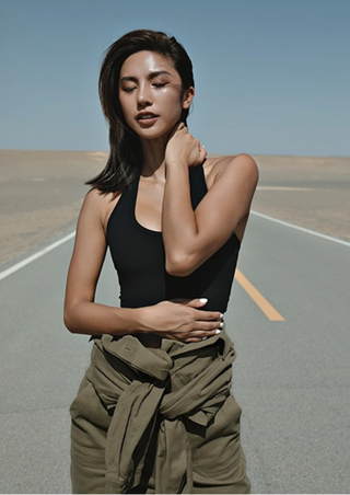 A confident woman stands on an empty road under a clear blue sky. She wears a black Halterneck Support Sports Bra and olive green pants with a matching jacket tied at her waist. One hand touches her neck while the other rests on her stomach, eyes closed and hair windswept.