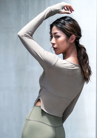A woman with long brown hair tied back wears the Curve Hugging Breathable Sleeves in taupe and olive green leggings. Posing confidently with her left arm raised and bent, she gazes to the side against a softly blurred neutral background.