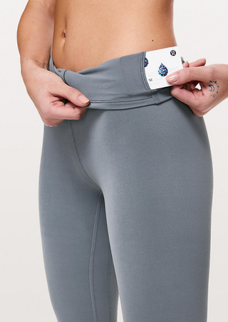 A person in fitted High-Waist Powercore Leggings in Lotus Gray pulls down the waistband to reveal a small pocket with part of a card inside. Their fingers, adorned with a heart tattoo, stand out against the plain, light background.