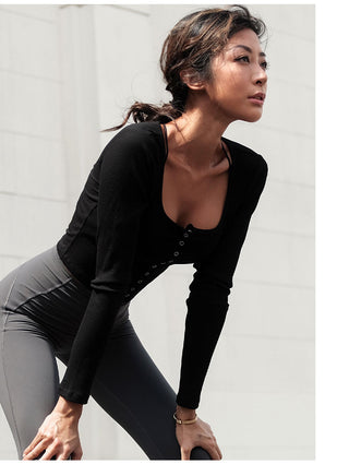 A woman with dark hair tied back, wearing the black Vibe Buttons Knitted Top and gray high-waisted leggings, leans forward with hands on her knees against a light-colored, textured wall. She has a focused expression and appears to be in an active or relaxed pose.