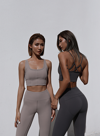 Two women in athletic wear stand against a white wall. The woman on the left is wearing a beige sports bra and leggings, while the woman on the right has her back partly turned, showcasing the Meta Crisscross Padded Top in gray with matching leggings. Both have medium-length hair.