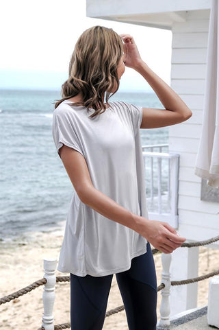 A woman with wavy, shoulder-length blonde hair stands on a beach in front of a white wooden structure. She is wearing the Love Street Style Top, a light gray, loose-fitting short-sleeve top, paired with dark pants. The ocean and sandy beach are visible in the background. She is facing slightly away and touching her hair with one hand.