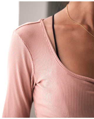 Close-up of a person wearing the Vibe Buttons Knitted Top in ribbed, light pink with long sleeves. The top reveals a black bra strap. A thin, gold chain necklace rests around their neck. The skin tone is light, and the background is out of focus with neutral colors.