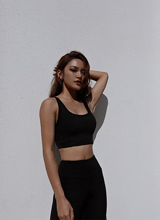 A woman with long brown hair stands against a plain white wall. She is wearing a black Meta Crisscross Padded Top and matching high-waisted leggings. Her left arm is raised with her hand resting behind her head, and she gazes towards the camera in a confident pose.