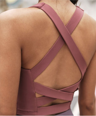 A woman is wearing a red Athleta Empower Sports Bra featuring an intricate criss-cross strap design on the back. The smooth, fitted fabric highlights the back details as she stands outdoors against a blurred urban backdrop.