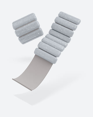 The Vibrant Bangles Set features light gray, cylindrical weights with a textured, speckled surface arranged in a 'V' shape. A beige strap extends diagonally underneath them against a plain white background.