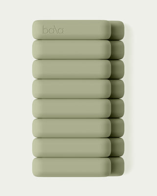 A stack of ten light olive green Vibrant Bangles, each rectangular with rounded corners, is arranged vertically on a pale background. The top bangle is subtly embossed with "bala.
