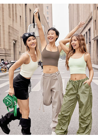 Three women are happily posing on an urban street. The first woman on the left wears a white crop top, black mini skirt, black knee-high boots, a black beret, and holds a green bag. The middle woman wears a FlexFit™ Ultimate Comfort Sports Bra and beige pants, raising her arms. The third woman wears a light green crop top and green baggy pants. All are smiling.