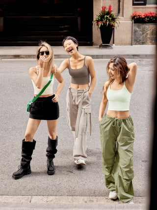 Three young women stand on a city street, smiling and looking carefree, each one sporting trendy, casual outfits. The woman on the left wears a white crop top, black mini skirt, chunky black boots, and sunglasses. The woman in the center pairs her FlexFit™ Ultimate Comfort Sports Bra with wide-legged pants for a chic yet comfy look. The woman on the right rocks a light green crop top and matching flowy pants.