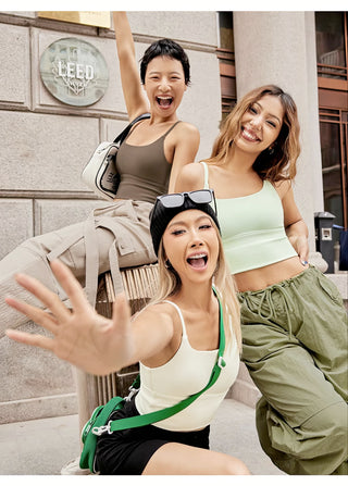 Three women are posing outdoors, smiling and looking cheerful. They are dressed in casual, sporty attire, with two wearing the FlexFit™ Ultimate Comfort Sports Bra and one in a tank top. One woman is playfully reaching towards the camera. A "Leed" sign is visible in the background, attached to a stone facade.