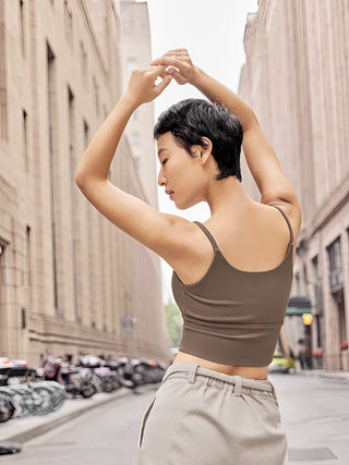 A person with short dark hair stands on a city street with their back toward the camera, wearing the FlexFit™ Ultimate Comfort Sports Bra and light-colored pants. Their arms are raised and crossed above their head. The street is flanked by tall buildings, and parked bicycles are visible in the background.