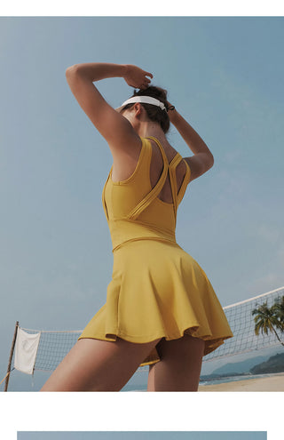 A woman stands on a beach with a volleyball net in the background. She is wearing an AeroFlex Athleisure Tennis Dress in yellow, along with a white visor, and is facing away from the camera with arms raised, hands behind her head. A palm tree and the ocean are visible in the distance under a clear blue sky.