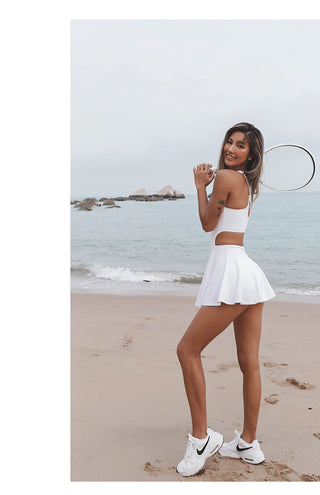 A woman is standing on a sandy beach near the water, smiling and holding a tennis racket. She is dressed in an AeroFlex Athleisure Tennis Dress and wearing white Nike sneakers. The background shows calm sea waves and a few rocks in the distance.