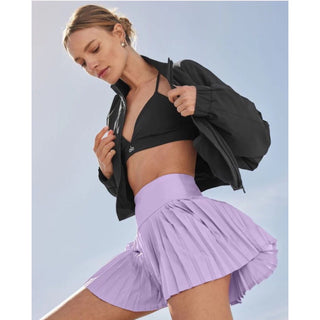 A woman stands against a clear blue sky, wearing a black sports bra and jacket, paired with the SerenityFlow™ Pleated Performance Skirt in lilac. She is looking down with a focused expression, her blonde hair pulled back, and she is accessorized with small hoop earrings.