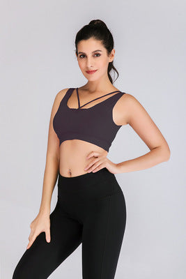 A woman with dark hair styled in a high ponytail is wearing the Ultimate Strappy-Back Sports Bra and high-waisted black leggings. She stands against a plain light gray background, posing confidently with one hand on her hip and the other resting by her side.