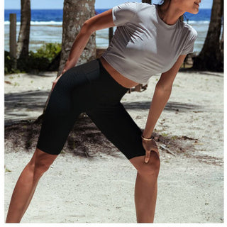 A woman wearing a grey short-sleeve crop top and High Waist Seamless Cycling Shorts in black is seen stretching outdoors near a beach. Her right knee is bent and her right hand is resting on her knee. The backdrop includes trees and the ocean with a blue sky, creating a sunny and calm scene.