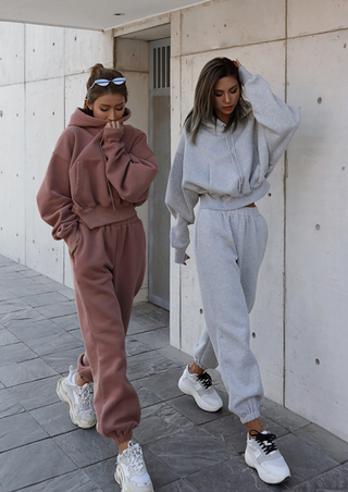 Two individuals stroll along a stone pavement beside a concrete wall. One is dressed in the Fashion Thick Tracksuit Women's set in brown, paired with white sneakers and sunglasses perched on their head. The other sports the same tracksuit in gray, also complemented by white sneakers. Both have their hands tucked into their pockets, exuding a relaxed vibe.