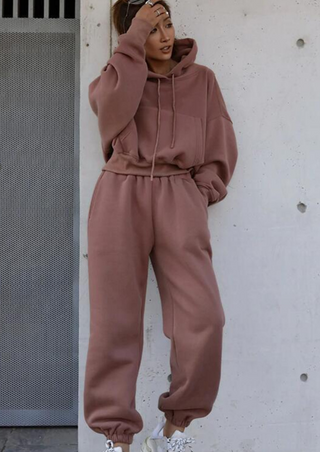 A person stands against a light-colored wall wearing the Fashion Thick Tracksuit Women in Bean Paste Powder. They have white sneakers, with their hair tied up. One hand rests on their head, and they appear relaxed and casual. A metal door is partially visible on the left.