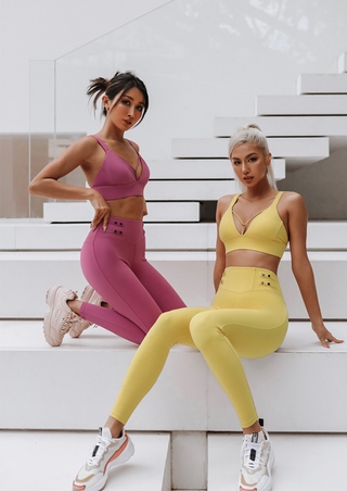 Two women wearing the Flow Y Gaudy Suit pose on white steps. The woman on the left sports a matching pink Flow Y Gaudy sports bra and leggings paired with sneakers, while the woman on the right wears a matching yellow Flow Y Gaudy sports bra and leggings with white sneakers. They are set against a modern, minimal backdrop featuring glass railings and stairs.