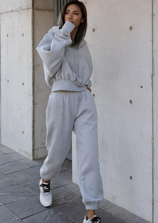 A person wearing the Fashion Thick Tracksuit Women in light grey stands against a concrete wall. They have on a hooded sweatshirt and matching sweatpants, paired with white and black sneakers. The individual is gazing to the side, with one hand near their face. The ground is paved with gray tiles.