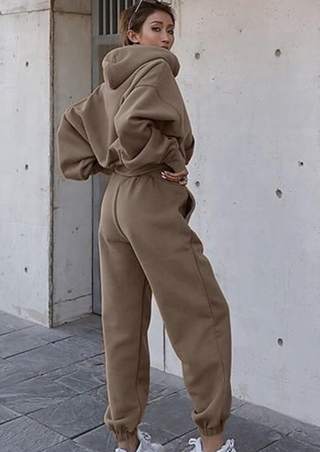 A person is standing outdoors wearing the Fashion Thick Tracksuit Women in apricot, which includes a loose-fitting hoodie and matching sweatpants. The pants are tucked into white sneakers as the individual poses sideways, glancing back over their shoulder. The setting is against a backdrop of a concrete wall and tile flooring.