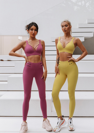 Two women pose confidently on white steps in their Flow Y Gaudy Suits. The woman on the left wears a matching fuchsia sports bra and leggings, paired with light pink sneakers. The woman on the right wears a matching yellow sports bra and leggings, paired with white sneakers. Both stand with hands on hips.