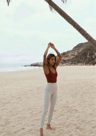 A woman stands barefoot on a sandy beach with her arms raised above her head. She is wearing a vibrant Radiant Bare Back Unitard, highlighting her silhouette against the backdrop. A palm tree is bent over her, and rocky hills are visible in the background. The sky is overcast, and the ocean waves are gentle.