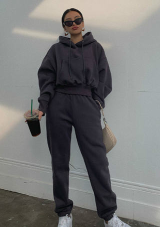 A person stands against a plain wall, dressed in the Fashion Thick Tracksuit Women set in Dark Grey. They hold a large iced coffee in one hand and a light beige handbag in the other. Their hair is pulled back, and they're accessorized with black sunglasses and white sneakers.