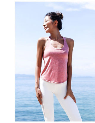 A woman stands in a relaxed pose by the ocean, smiling and looking upwards. She is wearing a pink "Tank top + Bra" and white leggings. Her hair is tied in a messy bun. The background features a clear blue sky and calm ocean waters, creating a peaceful and sunny atmosphere.