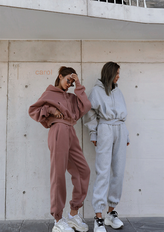 Two people stand against a concrete wall wearing cozy, oversized Fashion Thick Tracksuit Women sets. One is dressed in a dusty rose hue while the other sports a gray colorway. Both complement their outfits with white sneakers. The person in rose adjusts their sunglasses as the one in gray stands with hands casually tucked into their pockets.