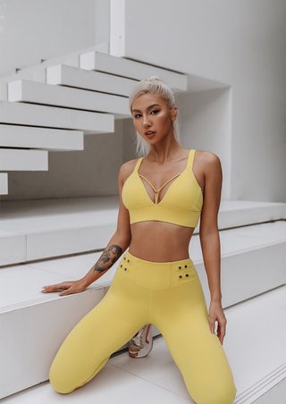 A woman with platinum blonde hair is kneeling on white stairs, dressed in the Flow Y Gaudy Suit—a vibrant yellow sports bra paired with matching high-waisted leggings. The setting appears modern and minimalistic with a monochromatic white background. She has a tattoo on her left arm and a focused expression.