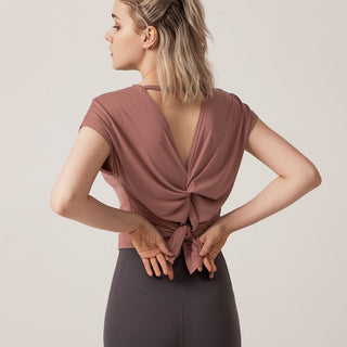 A woman with light hair tied back stands facing away, donning the Love Street Style Top, characterized by its mauve hue and deep V-back design. The top is elegantly tied in a bow at the waist. She pairs it with dark high-waisted leggings. The plain, light-colored background highlights her silhouette as she adjusts the bow on her top with her hands behind her.