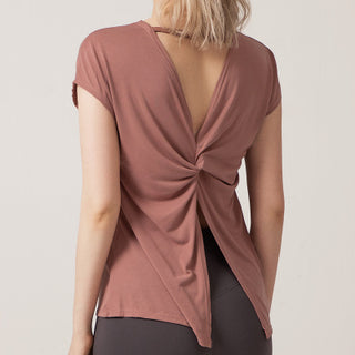 A woman with blonde hair is shown from the back, wearing the Love Street Style Top in old rose. The short-sleeve top features a twisted knot design in the center, creating a draped, open-back effect. She also wears dark leggings. The image background is plain and lightly colored.