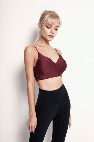 A person with blonde hair and bangs is wearing a red Silky Glow Sports Bra and black high-waisted leggings. They are standing against a plain white background, facing slightly to the side with their eyes closed. Their posture is relaxed, with one hand hanging down and the other not visible.