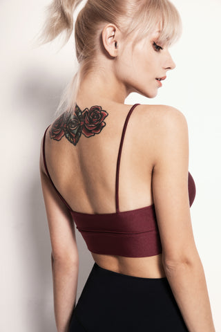 A person with light blonde hair tied in a ponytail is shown from the side, looking down. They wear a maroon Silky Glow Sports Bra with black bottoms. On their upper back, a detailed tattoo of red roses with green leaves is visible. The background is plain and light-colored.