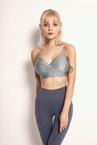 A woman with blonde hair, wearing the Silky Glow Sports Bra and blue leggings, stands against a neutral background. She has a tattoo on her right upper arm and her bangs frame her face. The woman looks directly at the camera with a neutral expression and her arms relaxed by her sides.