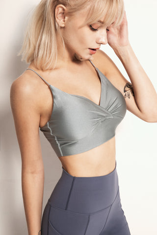 A woman with shoulder-length blonde hair is seen wearing a grey Silky Glow Sports Bra and high-waisted blue leggings. She has a tattoo on her right upper arm and is looking down while touching her hair. The background is a plain off-white wall.