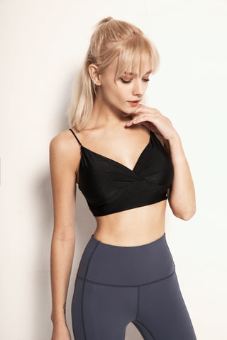 A woman with blonde hair in a ponytail stands against a plain background. She is wearing the Silky Glow Sports Bra in black and high-waisted gray leggings. Her right hand is raised to her chin, and she gazes downward with a thoughtful expression.