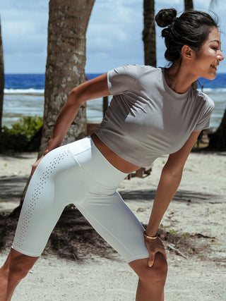 A woman in a gray crop top and High Waist Seamless Cycling Shorts bends sideways while smiling. She is outdoors, with a beach and trees visible in the background. Her hair is tied up, and she appears to be stretching or exercising.