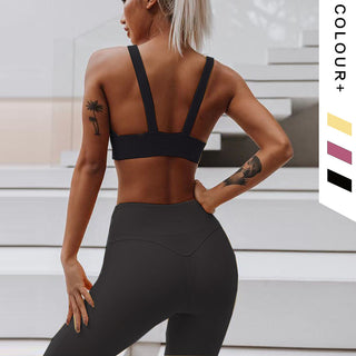 A woman with platinum blonde hair is wearing the Flow Y Gaudy Suit in black, standing with her back facing the camera. She has tattoos on her upper left arm and right forearm. The background features white stairs, and to the right are color swatches in purple, beige, and black labeled "COLOUR+".